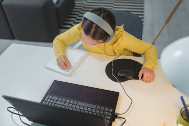 Kulaklık takan sevimli çocuk internetten öğrenmek için laptop kullanıyor. Ev okulu. Evde ödev yapan bir kız. Evde eğitim için yaşam tarzı konsepti. Sosyal uzaklık ve eğitim.