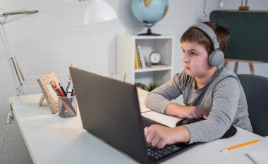 Kulaklık takan sevimli çocuk internetten öğrenmek için laptop kullanıyor. Ev okulu. Çocuk evde ödev yapıyor. Evde eğitim için yaşam tarzı kavramı
