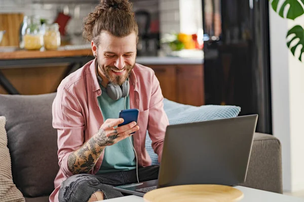 Young cheerful man with a sleeve tattoo using laptop computer and smartphone. Freelance entrepreneur working from home using banking apps.