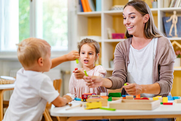 Kindergarten teacher playing together with children at colorful preschool classroom. Mother playing with children.