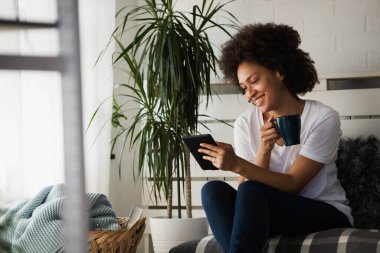 Genç Afrikalı Amerikalı kadın oturma odasında rahat bir koltukta oturuyor ve dijital tablet kullanıyor. 