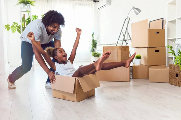 Father push cute little daughter sitting inside of carton boxes having fun riding in living room. Loan mortgage, housing improvement concept. Cheerful, happy african family enjoy relocation day.