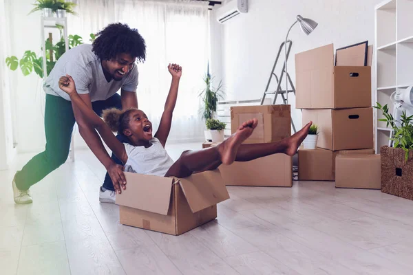 Father push cute little daughter sitting inside of carton boxes having fun riding in living room. Loan mortgage, housing improvement concept. Cheerful, happy african family enjoy relocation day.