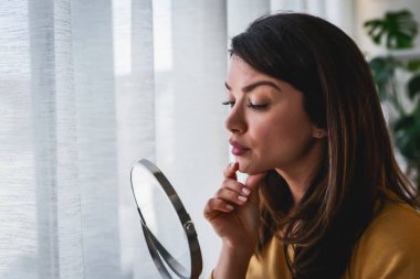 Evdeki aynada kendine bakan kadın büyük pencerenin yanında duruyor. Sivilceler konusunda endişeli.