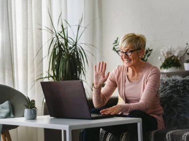 Güzel yaşlı kadın ailesiyle iletişim kurmak için dizüstü bilgisayar kullanıyor, internette sörf yapıyor, internetten alışveriş yapıyor, kahve içiyor.