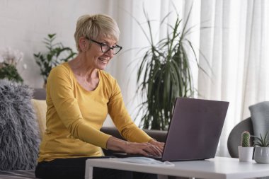 Güzel yaşlı kadın ailesiyle iletişim kurmak için dizüstü bilgisayar kullanıyor, internette sörf yapıyor, internetten alışveriş yapıyor, kahve içiyor.