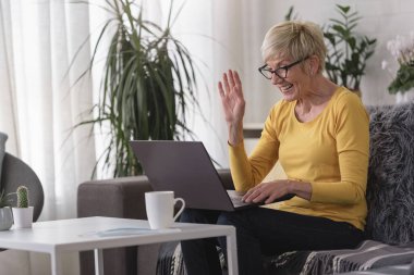 Güzel yaşlı kadın ailesiyle iletişim kurmak için dizüstü bilgisayar kullanıyor, internette sörf yapıyor, internetten alışveriş yapıyor, kahve içiyor.