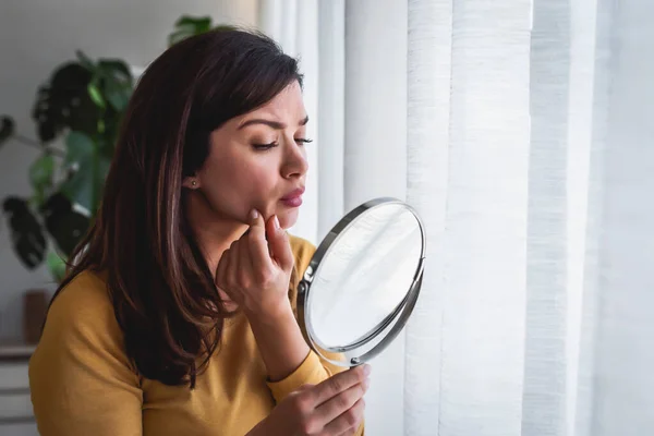Woman looking herself in the mirror at home standing by the big window. She is concerned about acne