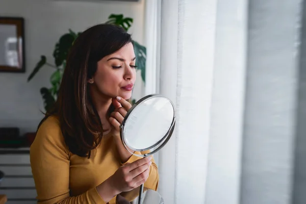 Woman looking herself in the mirror at home standing by the big window. She is concerned about acne