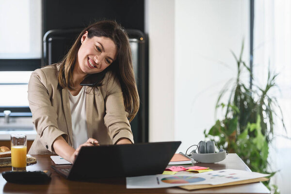 Young smiling businesswoman working from home sitting in front of laptop computer at home kitchen