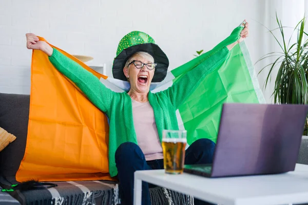 Old Irish lady watching the game , drinking beer, cheering for Ireland with an Irish flag and leprechaun hat. Winning a game!