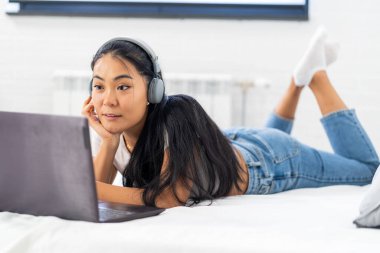 Asyalı bir öğrenci, evde internet üzerinden İngilizce öğrenen genç bir kadın. Kulaklık ve dizüstü bilgisayar kullanarak, online bir derse katılarak.
