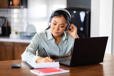 Çevrimiçi yabancı dil derslerine katılan genç Asyalı kadınlar. Kulaklıklı bir dizüstü bilgisayarın önünde oturmuş kursları dinliyor ve notlar alıyorsun..