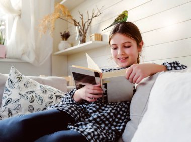 Evde kitap okuyan sevimli küçük kız, papağanıyla kanepede oturuyor.