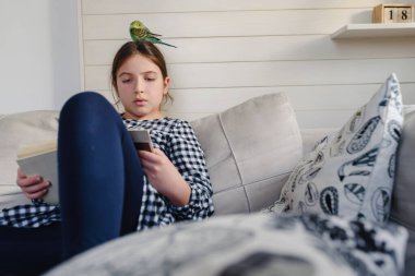 Evde kitap okuyan sevimli küçük kız, papağanıyla kanepede oturuyor.