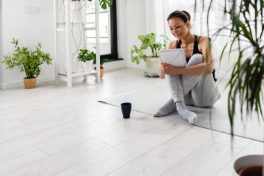 Defterine yazan genç bir kadın. Egzersiz sonrası evde, pencerenin yanında yoga minderinin üzerinde oturmak. Kişisel büyüme çözümleri