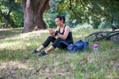 Bisiklete binmeye ara veren, parkta çimlerin üzerinde oturan ve cep telefonu kullanan genç bir kadın.