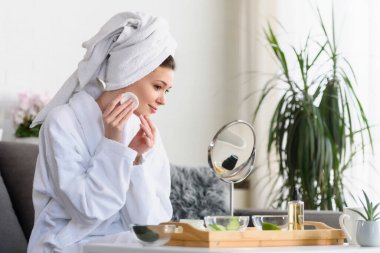 Bornoz ve havlu giymiş güzel genç bir kadın aynaya bakıyor, cilt bakımı konsepti