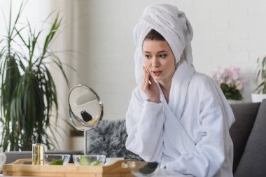 Bornoz ve havlu giymiş güzel genç bir kadın aynaya bakıyor, cilt bakımı konsepti