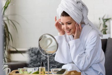 Bornoz ve havlu giymiş güzel genç bir kadın aynaya bakıyor, cilt bakımı konsepti