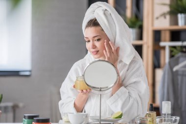 Bornoz ve havlu giymiş güzel genç bir kadın aynaya bakıyor, cilt bakımı konsepti