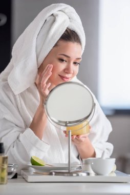 Bornoz ve havlu giymiş güzel genç bir kadın aynaya bakıyor, cilt bakımı konsepti