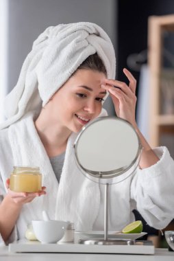 Bornoz ve havlu giymiş güzel genç bir kadın aynaya bakıyor, cilt bakımı konsepti