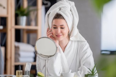 Bornoz ve havlu giymiş güzel genç bir kadın aynaya bakıyor, cilt bakımı konsepti