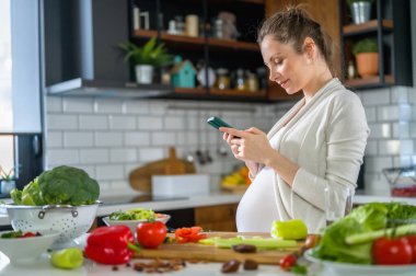 Elinde telefonla mutfakta bekleyen güzel hamile bir kadın.
