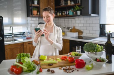 Elinde telefonla mutfakta bekleyen güzel hamile bir kadın.
