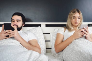 Genç çift yatakta uzanıyor ve cep telefonu kullanıyor.
