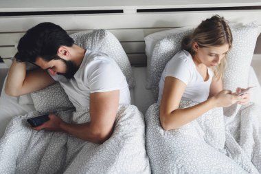 Genç çift yatakta uzanıyor ve cep telefonu kullanıyor.