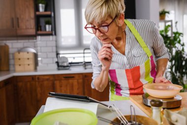 Yaşlı kadın ev mutfağında yemek pişiriyor. Tarifi bulmak için tablet bilgisayar kullanmak. İnternetin yardımıyla yemek yapan olgun bir kadın. Yaşlılar için modern teknoloji.