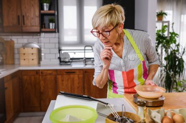 Yaşlı kadın ev mutfağında yemek pişiriyor. Tarifi bulmak için tablet bilgisayar kullanmak. İnternetin yardımıyla yemek yapan olgun bir kadın. Yaşlılar için modern teknoloji.
