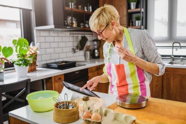 Yaşlı kadın ev mutfağında yemek pişiriyor. Tarifi bulmak için tablet bilgisayar kullanmak. İnternetin yardımıyla yemek yapan olgun bir kadın. Yaşlılar için modern teknoloji.