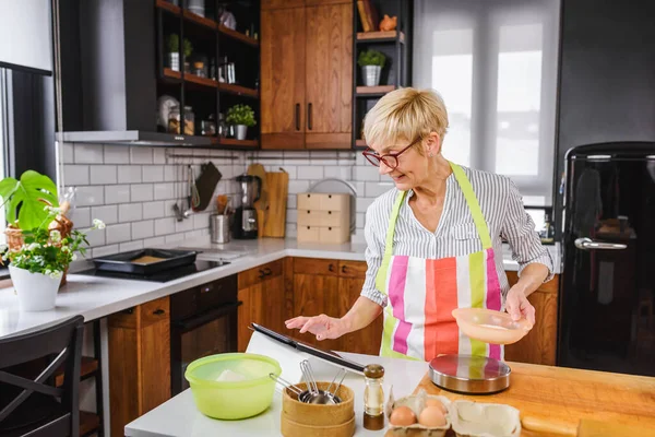 stock image Senior aged woman baking in home kitchen. Using tablet computer to find recipe. Mature woman cooking with the help of internet. Modern technology for older people.