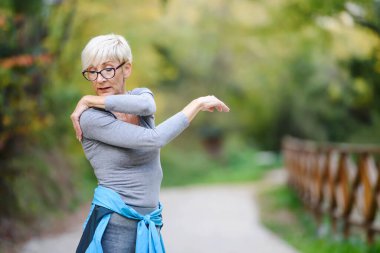 Gülümseyen yaşlı kadın parkta koşmadan önce geriniyor. Yaşlılar için spor faaliyetleri.