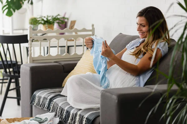 Hermosa Mujer Embarazada Mirando Alguna Linda Ropa Bebé Casa Mientras —  Fotos de Stock