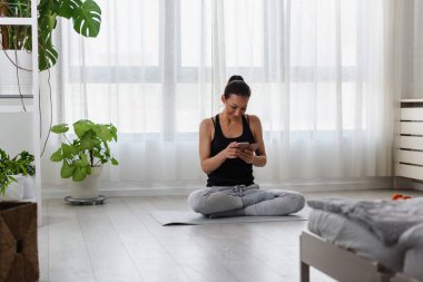 Genç ve sportif bir kadın evde egzersiz yaptıktan sonra akıllı telefon kullanıyor.