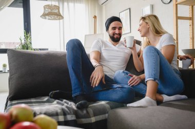 Genç hippi çift evdeki kanepede oturup konuşuyor ve kahve içiyor.