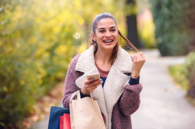 Güzel genç bir kadın alışveriş torbalarıyla parkta yürüyor.