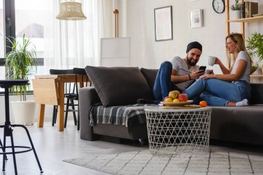 Genç ve mutlu bir adam telefonuyla evde oturmuş karısıyla kahve içiyor.