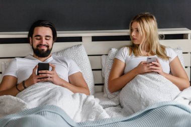 üzgün genç bir çift yatakta uzanıyor ve akıllı telefonlar kullanıyor.