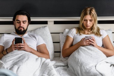 üzgün genç bir çift yatakta uzanıyor ve akıllı telefonlar kullanıyor.