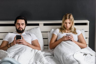 üzgün genç bir çift yatakta uzanıyor ve akıllı telefonlar kullanıyor.