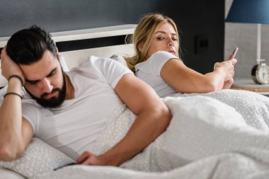 üzgün genç bir çift yatakta uzanıyor ve akıllı telefonlar kullanıyor.