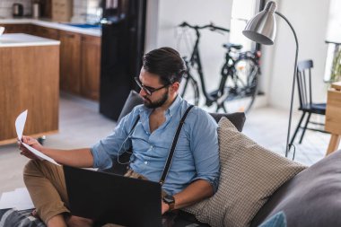 Genç adam dizüstü bilgisayar kullanıyor, evde çalışıyor.