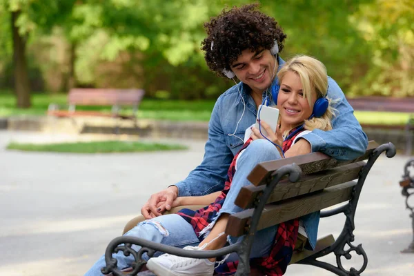 一对快乐的年轻夫妇坐在公园的长椅上听音乐 — 图库照片