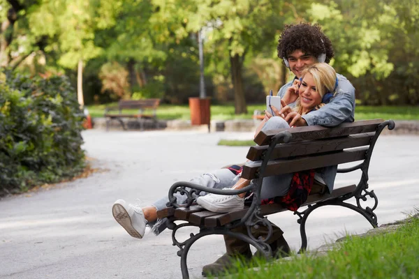 一对快乐的年轻夫妇坐在公园的长椅上听音乐 — 图库照片