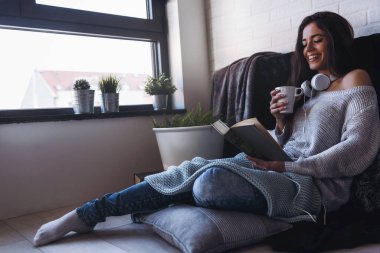Güzel genç bir kadın kitap okuyor, modern apartmanın penceresinin önünde eğleniyor..